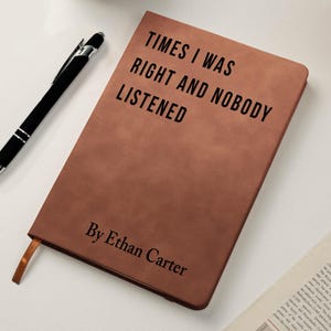 K&ouml;nnte beinhalten: Ein braunes Lederjournal mit dem Text "TIMES I WAS RIGHT AND NOBODY LISTENED" und "By Ethan Carter" in Schwarz gedruckt. Ein schwarzer Stift liegt neben dem Journal, was auf ein Schreibthema hindeutet.