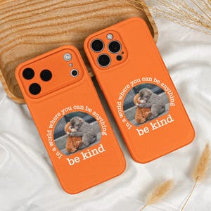May include: Two orange phone cases with a printed design. Each case features a circular image of a gray monkey hugging a brown teddy bear, with the words "In a world where you can be anything, be kind" in white.