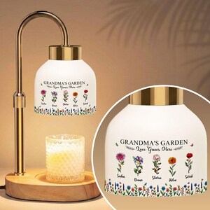 May include: A decorative lamp with a white shade featuring the text "GRANDMA'S GARDEN Love Grows Here" and floral illustrations. The lamp has a gold-colored metal frame and a wooden base. A lit candle sits below the shade.