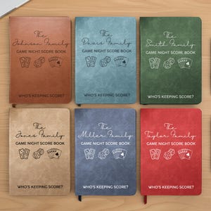May include: Six rectangular game night score books in brown, light blue, green, tan, dark blue, and red. Each cover features a family name, the text GAME NIGHT SCORE BOOK, illustrations of dominoes, dice, and playing cards, and WHO'S KEEPING SCORE?