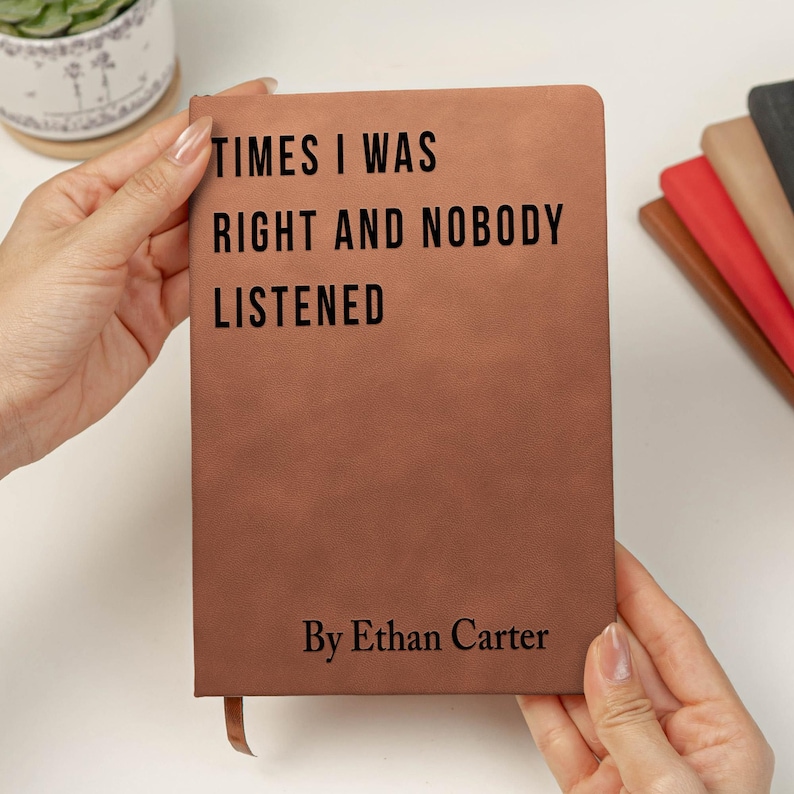 K&ouml;nnte beinhalten: Ein braunes Lederjournal, das von H&auml;nden gehalten wird, mit dem Text "TIMES I WAS RIGHT AND NOBODY LISTENED" und "By Ethan Carter" in Schwarz gedruckt. Ein Lesezeichen aus Band ist sichtbar. Weitere Journale in verschiedenen Farben sind im Hintergrund.