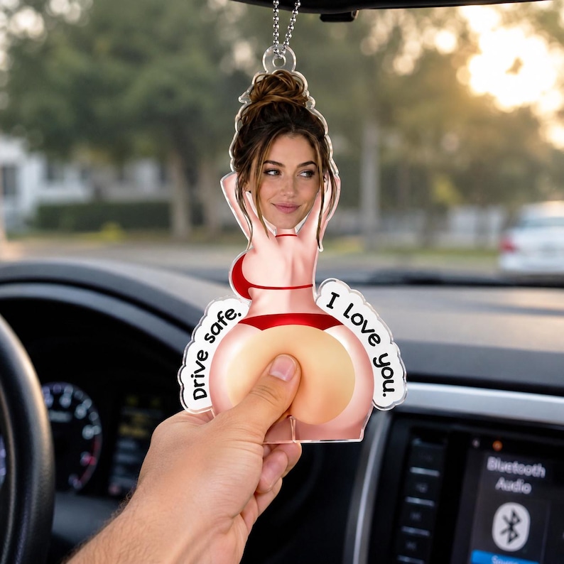 May include: A clear acrylic car air freshener ornament featuring a graphic illustration of a person in a red bikini with a photo-realistic face, accompanied by the printed text Drive safe. I Love you. hanging from a silver ball chain.
