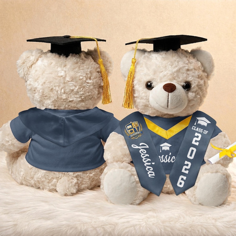 K&ouml;nnte beinhalten: Zwei cremefarbene Teddyb&auml;ren mit Abschlusskappe und -robe. Einer der B&auml;ren hat eine Sch&auml;rpe mit "Jessica" und "Class of 2026". Die B&auml;ren tragen dunkelblaue Roben und Kappen mit goldenen Quasten.