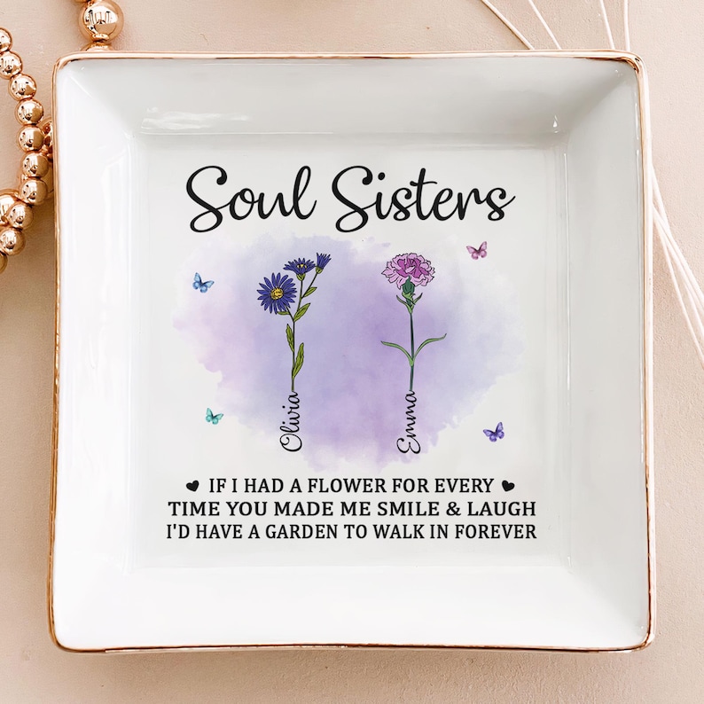 May include: White ceramic trinket tray with a gold rim. The tray features a watercolor design of two flowers with the text "Soul Sisters" above the flowers. The names "Olivia" and "Emma" are written below the flowers. The text "If I had a flower for every time you made me smile & laugh I'd have a garden to walk in forever" is written below the flowers.