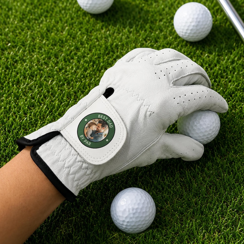 K&ouml;nnte beinhalten: Ein wei&szlig;er Golfhandschuh aus Leder mit einem kreisf&ouml;rmigen Aufn&auml;her am Handgelenkriemen. Der Aufn&auml;her ist dunkelgr&uuml;n mit wei&szlig;em Rand und zeigt ein Foto einer Person, die ein Kind h&auml;lt, mit dem in Wei&szlig; gedruckten Text BEST DAD BY PAR.