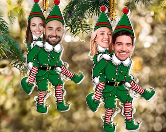 May include: Two Christmas ornaments featuring a couple dressed as elves. The elves are wearing green and red striped pants, green hats, and green jackets with white trim. The couple's faces are visible through the elf costumes. The ornaments are hanging from a Christmas tree.