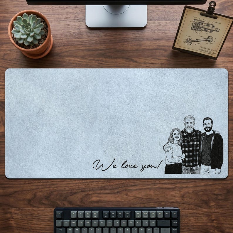 May include: A light blue rectangular mouse pad with a family portrait and the handwritten text "We love you!". The portrait is a black and white sketch of three people. A succulent plant in a terracotta pot is visible.