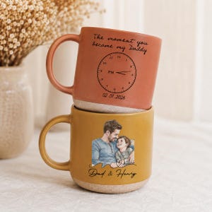 May include: Two ceramic mugs stacked on top of each other. The top mug is orange with the text "The moment you became my Daddy" and a clock design. The bottom mug is yellow with a photo of a father and child and the text "Dad & Henry".