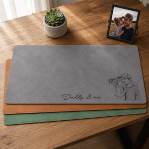May include: Three rectangular desk mats in gray, brown, and green are stacked on a wooden surface. The top gray mat has the text "Daddy & me" and a sketch of a father and child. A framed photo and a potted succulent are also present.