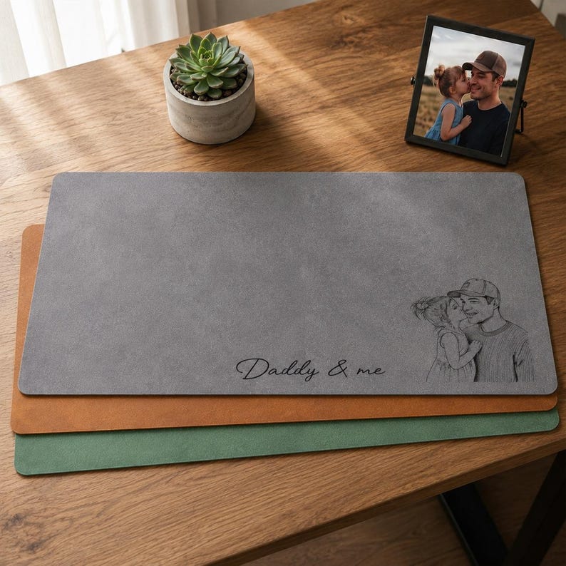 May include: Three rectangular desk mats in gray, brown, and green are stacked on a wooden surface. The top gray mat has the text "Daddy & me" and a sketch of a father and child. A framed photo and a potted succulent are also present.