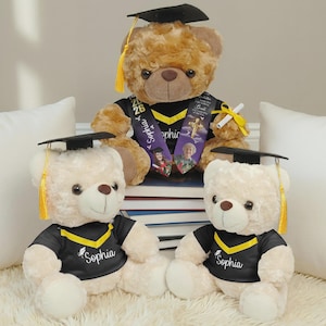 May include: Three plush teddy bears wearing black graduation caps with yellow tassels and black gowns with yellow V-neck trim. The two smaller bears have the name Sophia printed in white. The larger bear wears a sash with the year 2026.