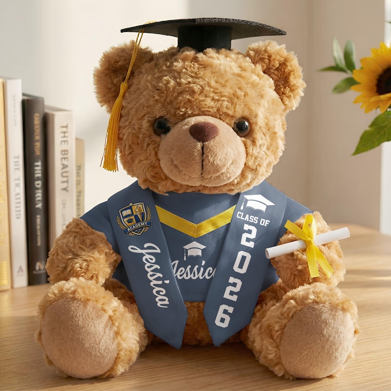 K&ouml;nnte beinhalten: Ein hellbrauner Teddyb&auml;r mit Abschlusskappe und blauem Sch&auml;rpe mit dem Text "Class of 2026" und dem Namen "Jessica". Der B&auml;r hat eine dunkelbraune Nase und schwarze Augen. Er h&auml;lt ein aufgerolltes Diplom.