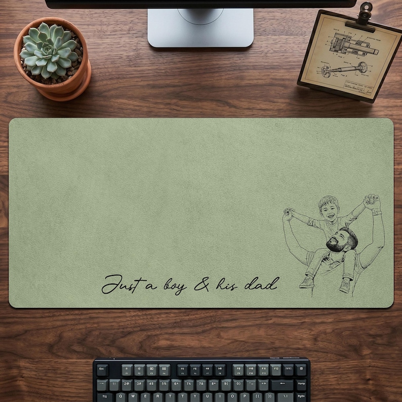 May include: Sage green rectangular desk mat with a black and white illustration of a father and son. The text "Just a boy & his dad" is written in cursive. A keyboard and computer monitor are visible.