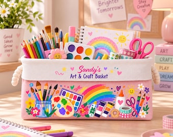 May include: A rectangular pink fabric storage basket with rope handles, labelled Sandy's Art & Craft Basket. The exterior features colourful illustrations of paintbrushes, a palette, a rainbow, and glue. It is filled with art supplies including coloured pencils, paint sets, and scissors.