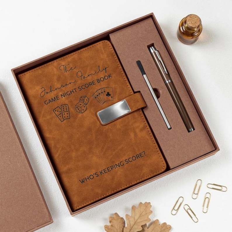 May include: A brown faux leather game night score book in a gift box with a metal pen, a stylus, and a small glass bottle. The cover features the text The Johnson Family GAME NIGHT SCORE BOOK, illustrations of dice and cards, and WHO'S KEEPING SCORE?
