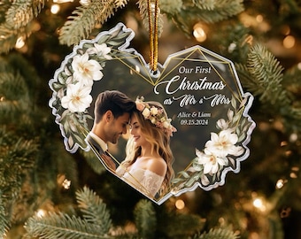 May include: Clear heart-shaped Christmas ornament with a photo of a couple, surrounded by white flowers and green leaves. Text reads "Our First Christmas as Mr & Mrs" with names and a date. Gold hanging string.