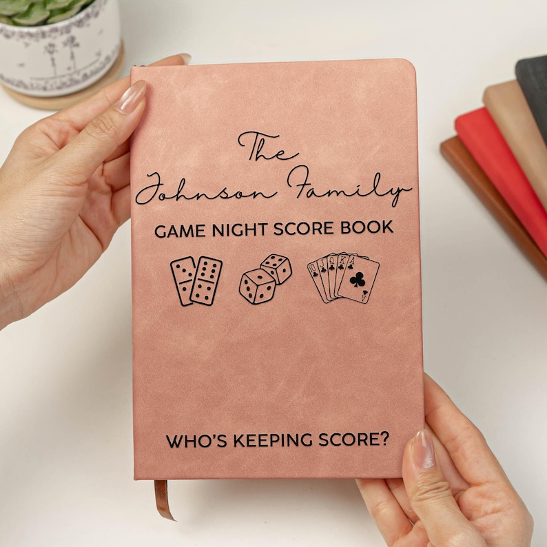 May include: A dusty rose-colored score book with black text reading The Johnson Family GAME NIGHT SCORE BOOK and WHO'S KEEPING SCORE? The cover features black line art illustrations of two dominoes, two dice, and a hand of five playing cards.