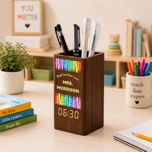 Könnte beinhalten: Ein rechteckiger Stiftehalter aus dunklem Holz mit einer Digitaluhr, die 06:30 anzeigt. Die Vorderseite ist mit einer bunten Buntstiftgrafik, dem kursiven Text "best teacher ever" und dem Namen "MRS. MORRISON" in gelben Großbuchstaben bedruckt.
