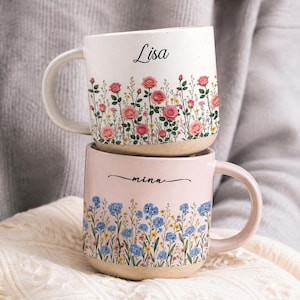 May include: Two ceramic mugs stacked vertically. The top mug is off-white with pink roses and the name Lisa in black script. The bottom mug is light pink with blue wildflowers and the name mina in black script. Both have a beige base.