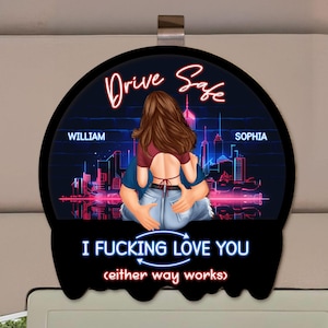 Könnte beinhalten: Schwarzer Autosonnenblende mit einem sich umarmenden Paar vor einer Skyline. Die Worte "Drive Safe" und "I Fucking Love You" sind im Neonstil dargestellt, zusammen mit den Namen "William" und "Sophia".