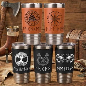 May include: Five stainless steel tumblers with leather-like sleeves. The top two are tan with black Viking symbols and runes. The bottom three are black with white designs featuring a tree of life, a compass, and two birds with runic inscriptions.