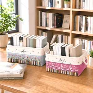 May include: Two rectangular fabric storage bins with rope handles, each filled with books. The left bin features the name Sophia, a floral print, and a book spine pattern. The right bin features the name Amelia, a pink floral print, and book spines.