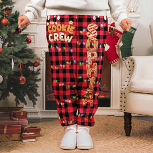 May include: Red and black plaid pajama pants with the text "THE COOKIE CREW" and the name "SOPHIA" in gingerbread cookie lettering. The pants are decorated with colorful string lights, perfect for the holiday season.