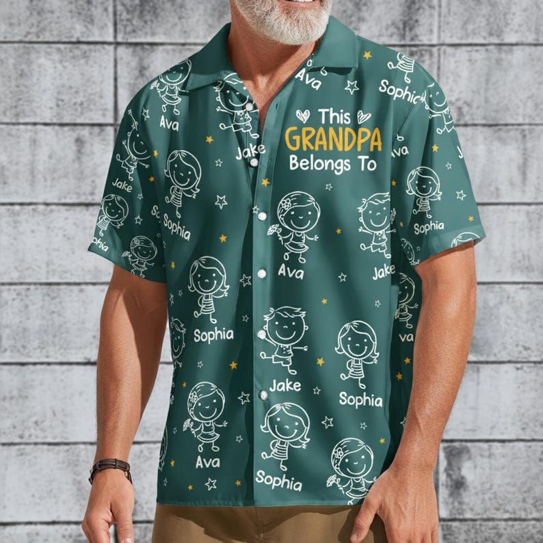 Peut inclure: Chemise boutonn&eacute;e bleu-vert avec un motif r&eacute;p&eacute;t&eacute; de personnages enfantins, d'&eacute;toiles et de noms. Le texte "This Grandpa Belongs To" est au centre. Les noms "Ava", "Jake" et "Sophia" sont visibles.