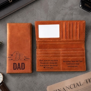 May include: A brown leather wallet, open and closed, with the word "DAD" and a fist bump design. The open wallet has card slots and a clear ID window. Text reads "Dad, you are my hero. Thanks for being the best dad in the world I love you."