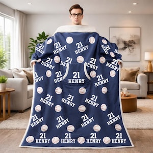 Könnte beinhalten: Eine marineblaue Kapuzen-Decke mit weißen Baseballs und dem Namen "HENRY" und der Nummer "21" bedruckt. Die Decke hat ein weißes Sherpa-Futter und eine große Fronttasche. Ein gemütlicher Artikel.