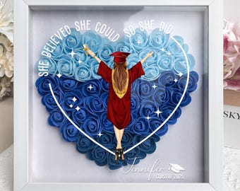 May include: A white frame with a heart-shaped arrangement of blue and light blue artificial roses. A graduate in a red cap and gown stands in the center of the heart. The text "She believed she could so she did" is written around the heart. The name "Jennifer" and "Class of 2025" are written below the heart.