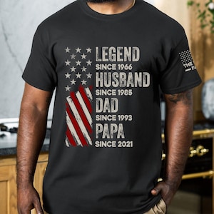 May include: Black t-shirt featuring an American flag design with text that reads "LEGEND SINCE 1966, HUSBAND SINCE 1985, DAD SINCE 1993, PAPA SINCE 2021." The shirt has a distressed, vintage look.