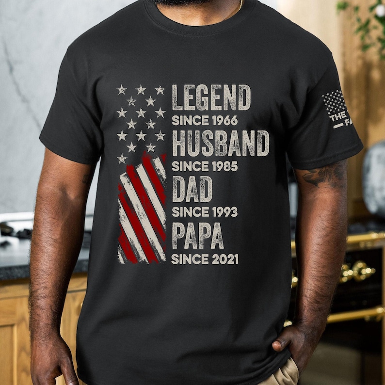 May include: Black t-shirt featuring an American flag design with text that reads "LEGEND SINCE 1966, HUSBAND SINCE 1985, DAD SINCE 1993, PAPA SINCE 2021." The shirt has a distressed, vintage look.