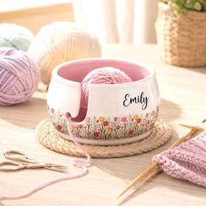 Könnte beinhalten: Eine weiße Keramik-Garnschale mit rosa Innenseite, verziert mit einem bunten Blumenmuster aus Tulpen und Wildblumen am Boden. Der Name Emily ist in schwarzer Schreibschrift auf der Seite aufgedruckt. Ein rosa Garnknäuel liegt in der Schale.