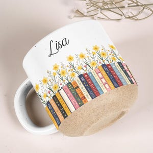 May include: A white ceramic mug featuring a printed design of colorful book spines arranged in a row, topped with yellow daffodil flowers and green stems. The name Lisa is printed in black script above the books. The base has a speckled tan texture.