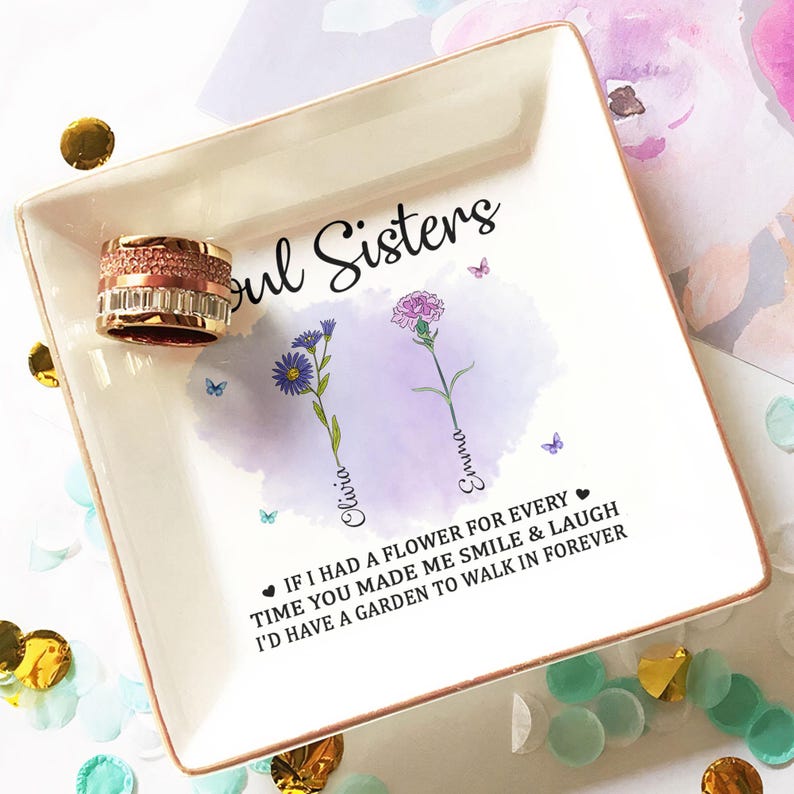 May include: White ceramic trinket tray with a gold rim. The tray features a watercolor design of two flowers with the names "Olivia" and "Emma" written next to them. The text "Soul Sisters" is at the top of the tray. The text "If I had a flower for every time you made me smile & laugh I'd have a garden to walk in forever" is at the bottom of the tray.