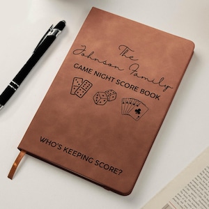 May include: A brown rectangular notebook with a black pen. The cover features the text The Johnson Family GAME NIGHT SCORE BOOK, illustrations of dominoes, dice, and playing cards, and the text WHO'S KEEPING SCORE? at the bottom.