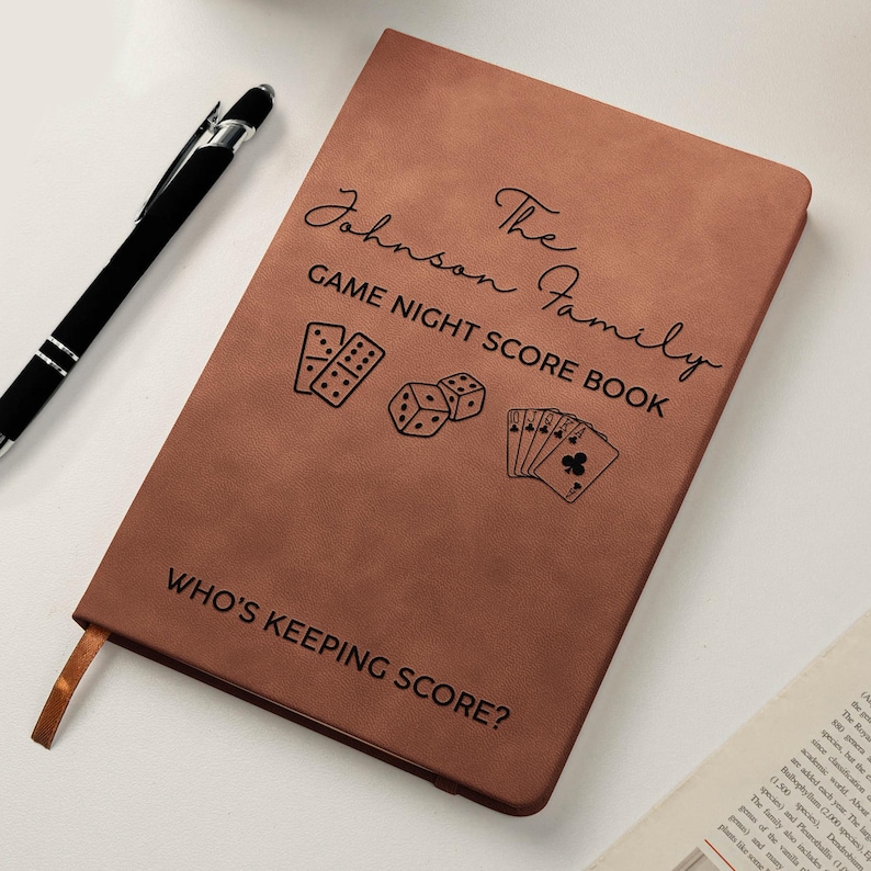 May include: A brown rectangular notebook with a black pen. The cover features the text The Johnson Family GAME NIGHT SCORE BOOK, illustrations of dominoes, dice, and playing cards, and the text WHO'S KEEPING SCORE? at the bottom.