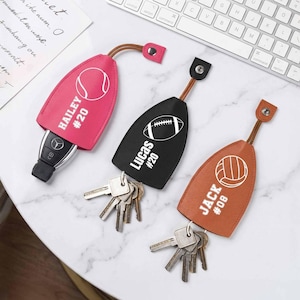 May include: Three personalized keychains in pink, black, and brown. Each keychain features a sports ball graphic: tennis, football, and volleyball. The names "Hailey," "Lucas," and "Jack" are printed with numbers.