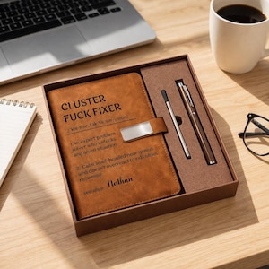 Könnte beinhalten: Ein braunes Leder-Notizbuch- und Stift-Set in einer Geschenkbox. Das Notizbuch trägt den Text "CLUSTER FUCK FIXER" und eine Definition. Ein silberner Stift und ein brauner Stift sind enthalten. Das Set befindet sich auf einer Holzoberfläche.