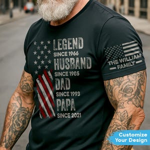 May include: Black t-shirt with a distressed American flag design and text. The text includes "LEGEND SINCE 1966", "HUSBAND SINCE 1985", "DAD SINCE 1993", and "PAPA SINCE 2021". The sleeve features an American flag and "THE WILLIAM FAMILY".