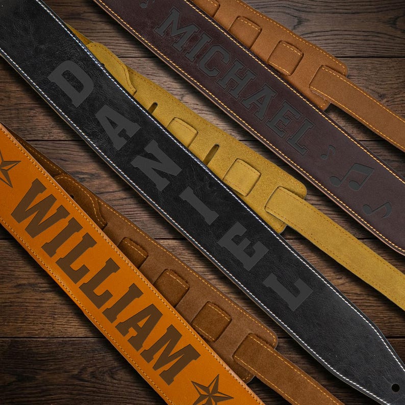 May include: Three guitar straps featuring personalized names and decorative elements. One strap is tan with the name WILLIAM and a star, one is black with the name DANIEL, and one is dark brown with the name MICHAEL and musical note symbols.