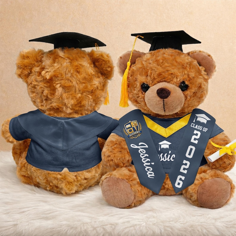 K&ouml;nnte beinhalten: Zwei braune Teddyb&auml;ren mit Abschlusskappen und -roben. Einer der B&auml;ren hat eine Sch&auml;rpe mit "Jessica" und "Class of 2026". Sie befinden sich auf einer wei&szlig;en, flauschigen Oberfl&auml;che.