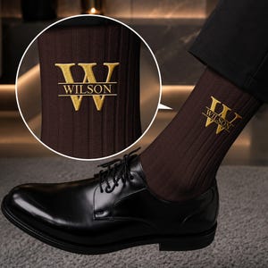 May include: A dark brown ribbed sock featuring an embroidered gold-colored letter W with the text WILSON centered horizontally across the letter. The sock is paired with a black leather dress shoe with black laces and a low heel.