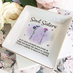 May include: White ceramic trinket dish with a gold rim. The dish features a watercolor design of two flowers with the names Olivia and Emma written on the stems. The text reads "Soul Sisters" and "If I had a flower for every time you made me smile & laugh I'd have a garden to walk in forever."