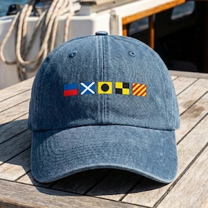 Peut inclure: Casquette de baseball en denim bleu avec des drapeaux de signalisation nautiques brodés. Les drapeaux sont rouges, blancs, bleus, noirs, jaunes et oranges. La casquette est posée sur une surface en bois.