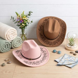 Pode incluir: Um chap&eacute;u de cowboy rosa claro com uma faixa tran&ccedil;ada escura e gravuras florais, com o nome Sophia, ao lado de um chap&eacute;u de cowboy bege com gravuras florais e o nome Emily, duas mantas enroladas, um vaso de flores e um pano estampado.