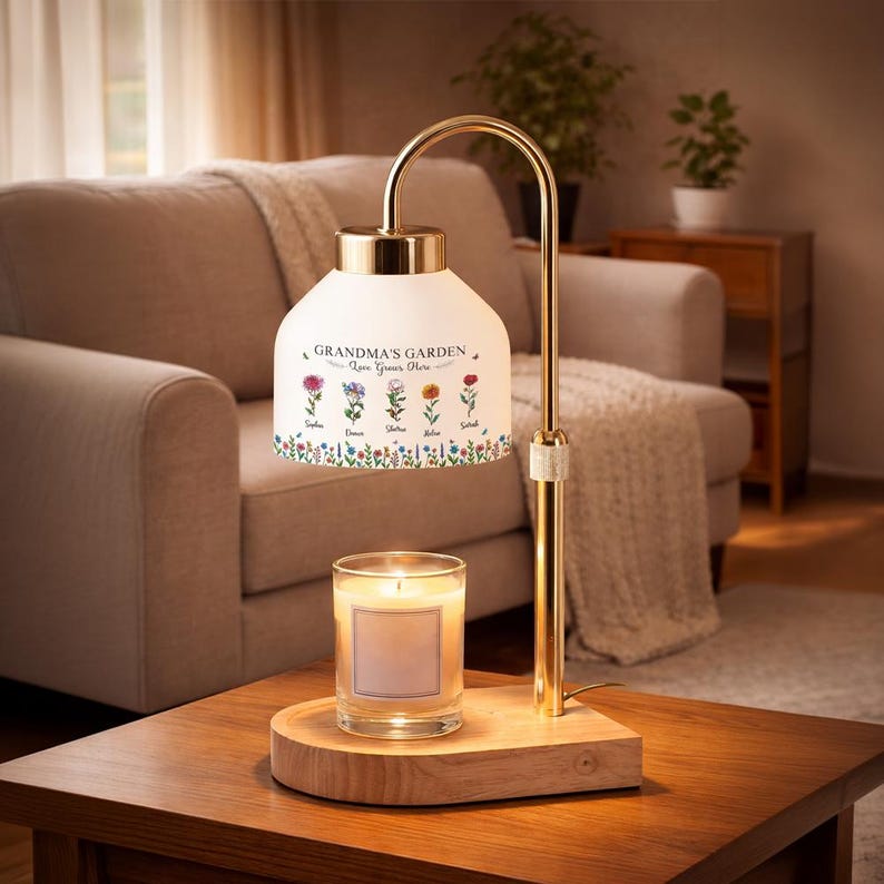 May include: A gold-toned lamp with a white shade featuring the text "Grandma's Garden" and floral illustrations. A lit candle sits on a wooden base below the lamp. The lamp is on a wooden table.