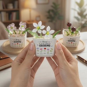 May include: Three small, square ceramic pots with glass flower arrangements. Each pot is white with the words "Blessed to be called GRANDMA" and a row of colorful flower illustrations. The flowers in the pots are clear, pink, and red.