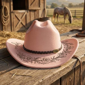 Pode incluir: Um chap&eacute;u de cowboy rosa claro com uma faixa tran&ccedil;ada castanho-escura e um pequeno emblema met&aacute;lico oval. A aba &eacute; decorada com ilustra&ccedil;&otilde;es de linhas pretas de motivos florais e o nome Sophia escrito em letra cursiva na lateral.